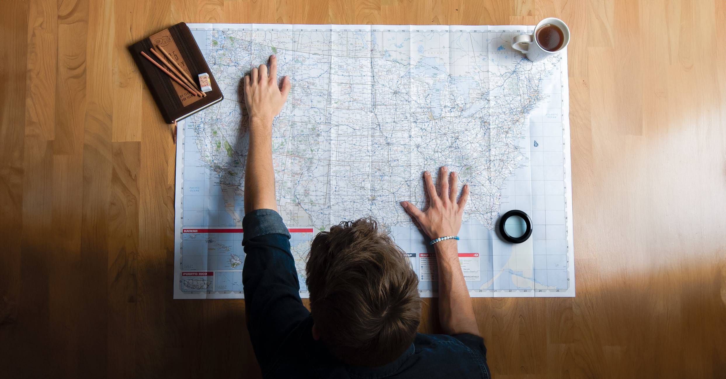person stands over map of the United States, cup of coffee, magnifier and journal with pencils laid out on table