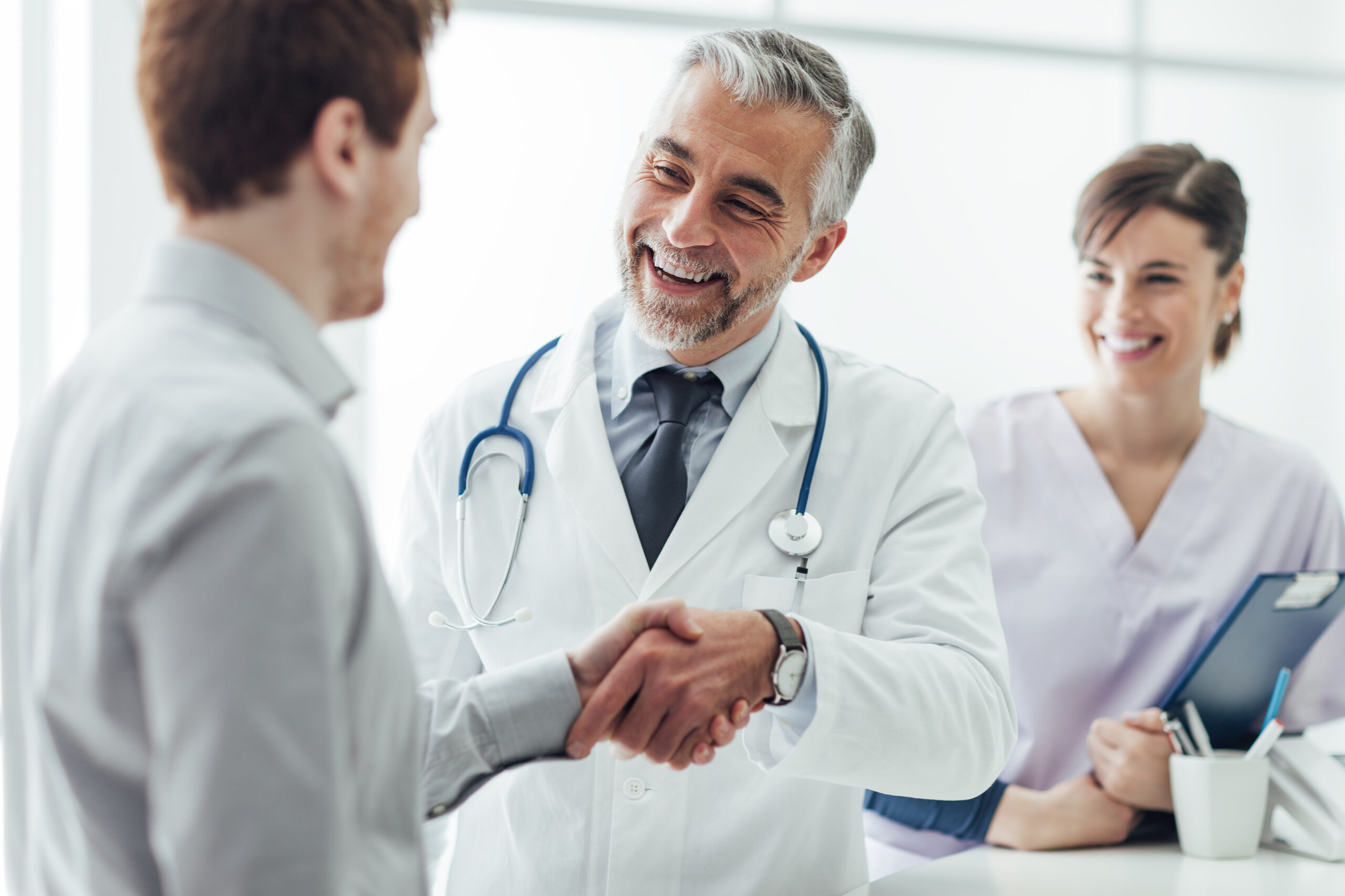 Mature male doctor smiling and shaking hands with patient while female nurse stands behind him