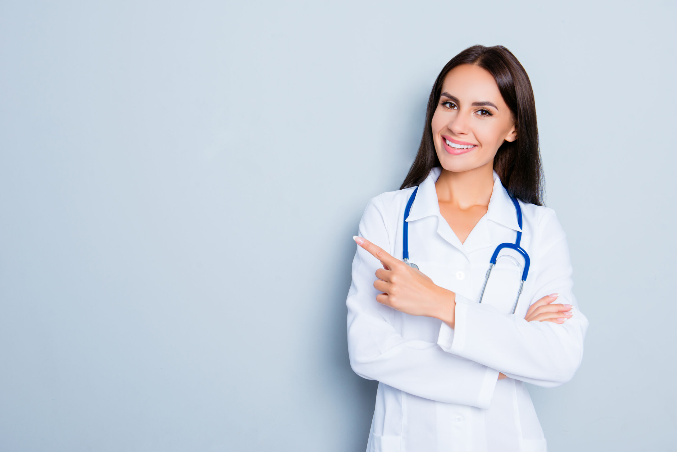 Female doctor wearing blue stethoscope, pointing her index finger past her shoulder. Light blue background.