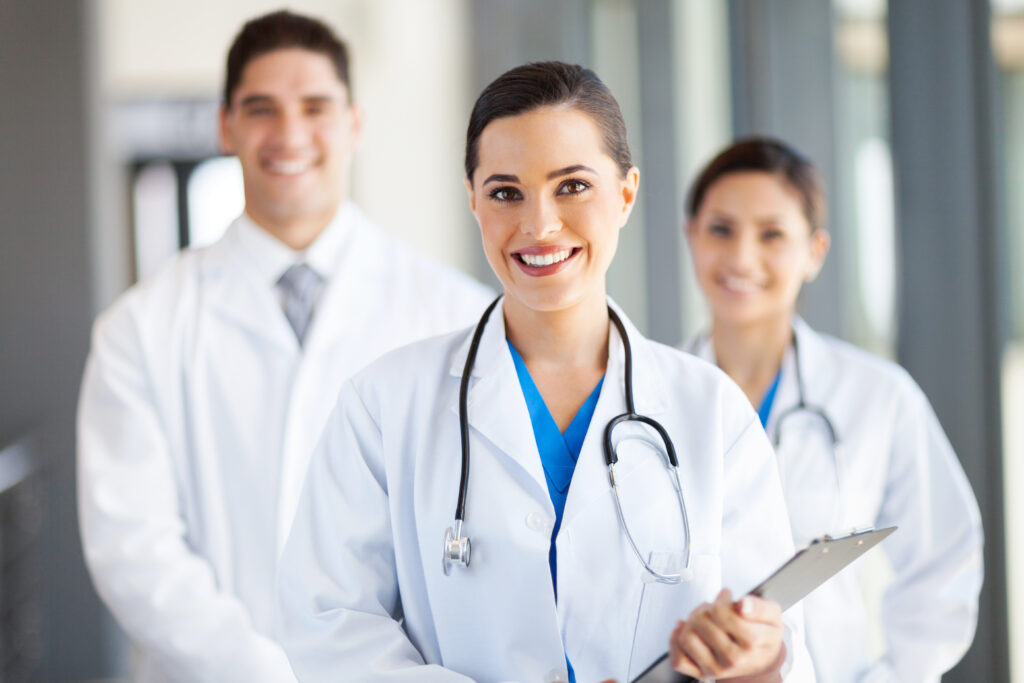 Young female doctor holding a clipboard smiles at camera while two other medical professional in lab coats stand behind her