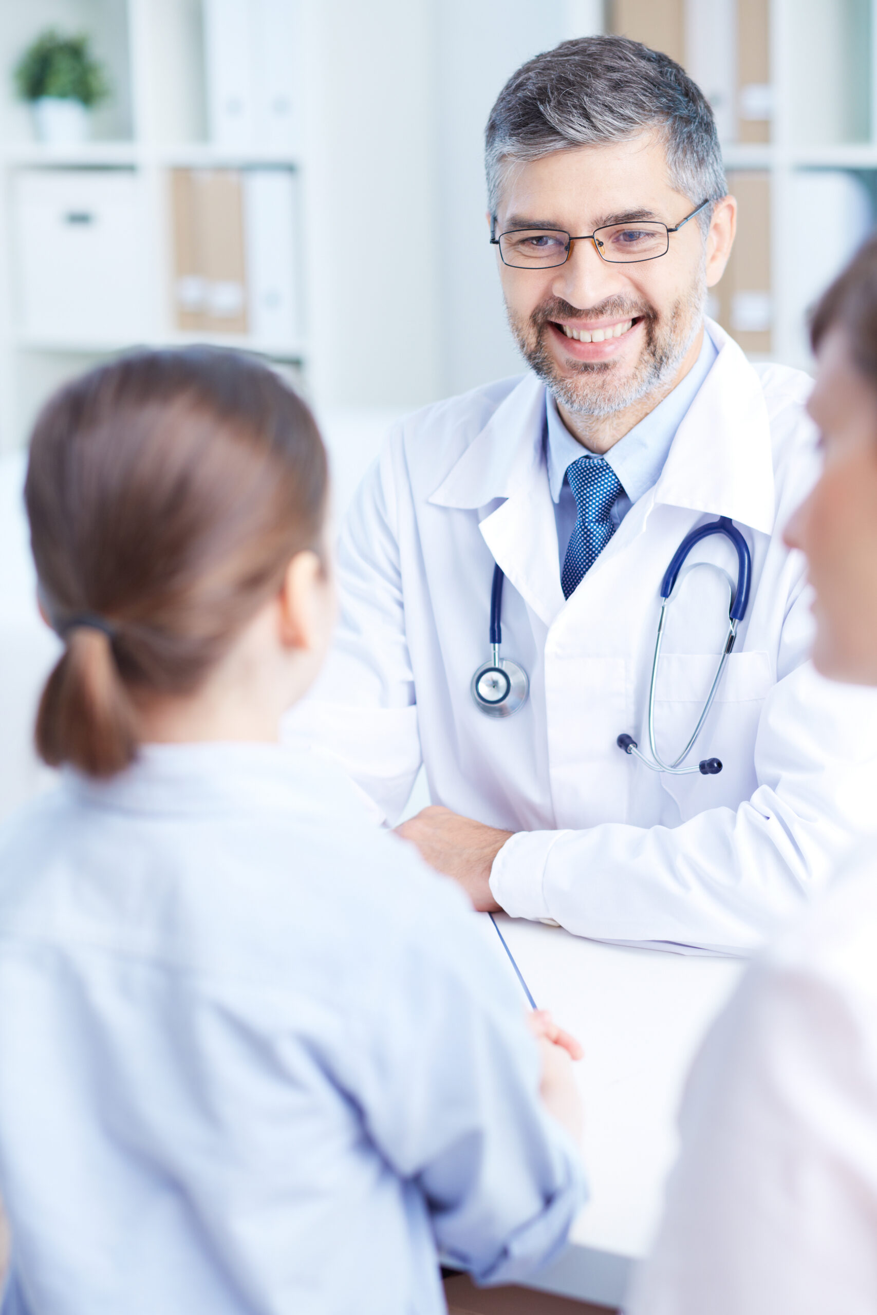 Mature medical doctor with grey hair stands at counter while smiling at young patient