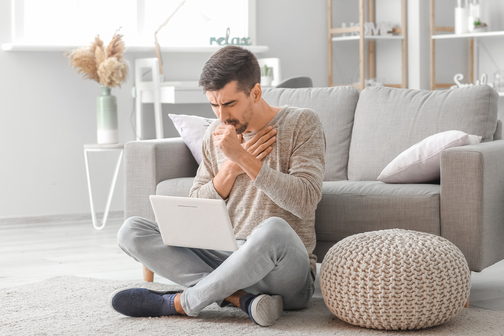 Man sitting on floor in grey living room holding small laptop and coughing